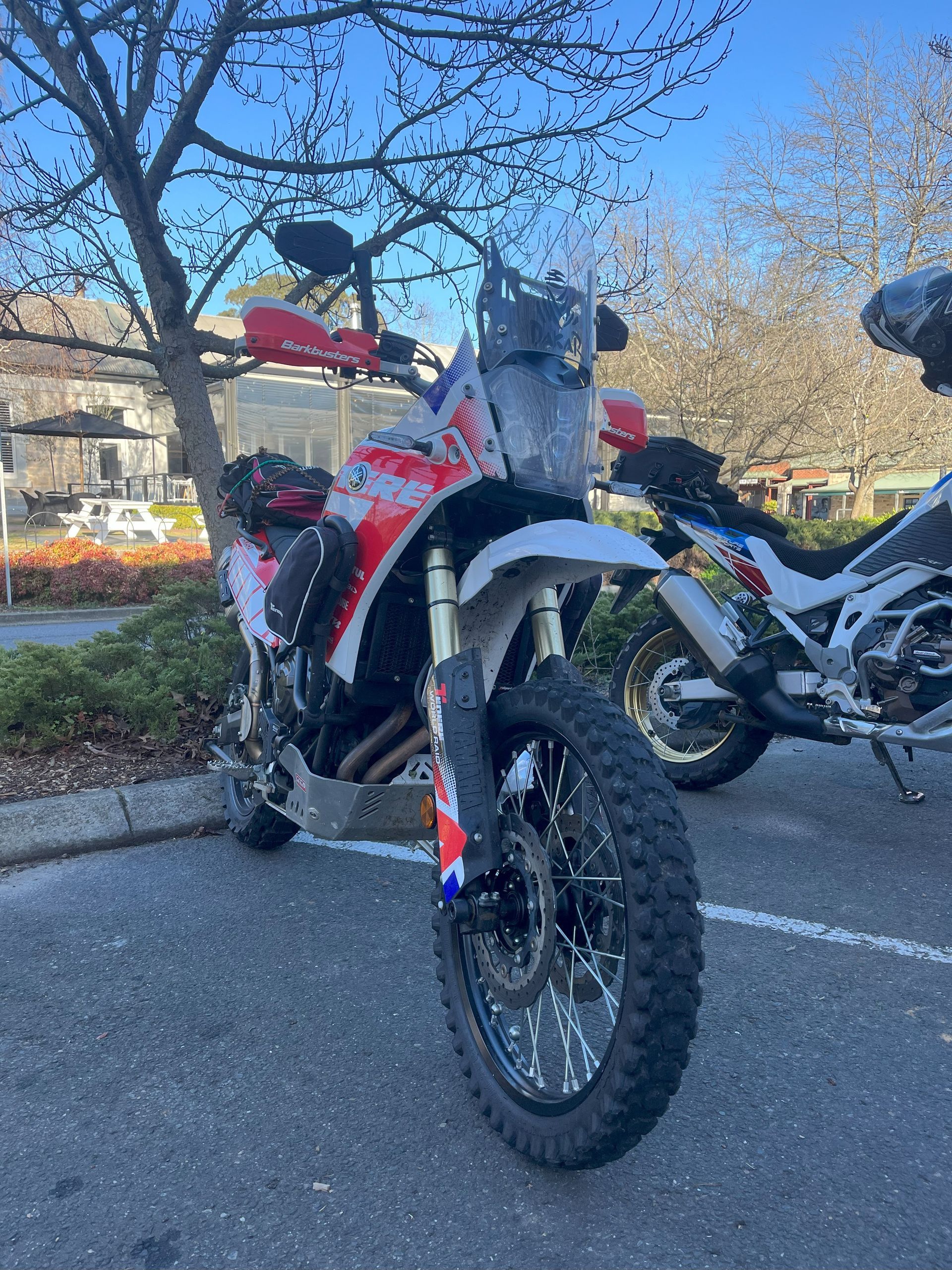 Two off-road motorcycles parked in a lot, one red-and-white bike in the foreground under trees.
