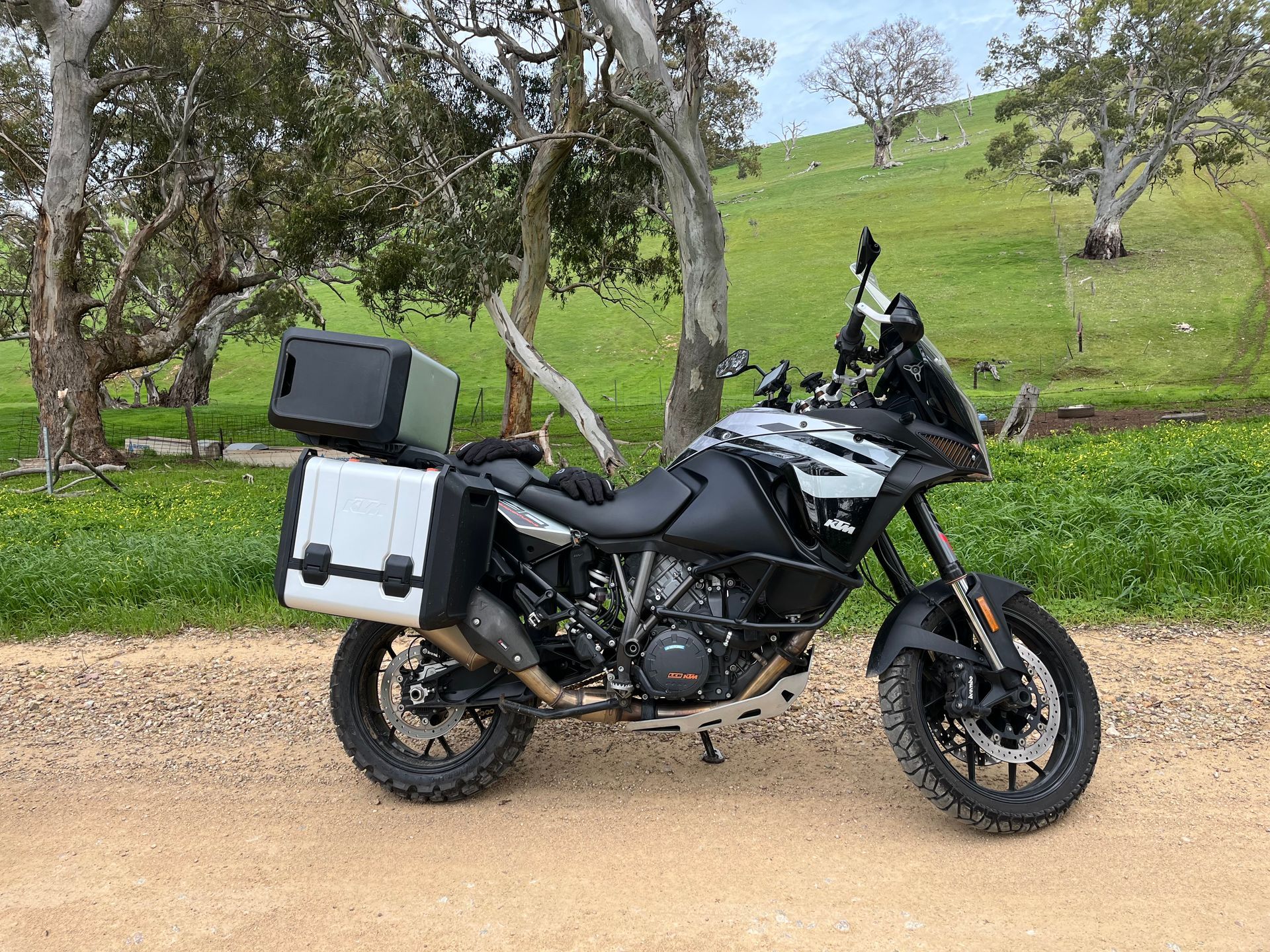 Black adventure motorcycle with panniers parked on a dirt path in a grassy, tree-lined field.