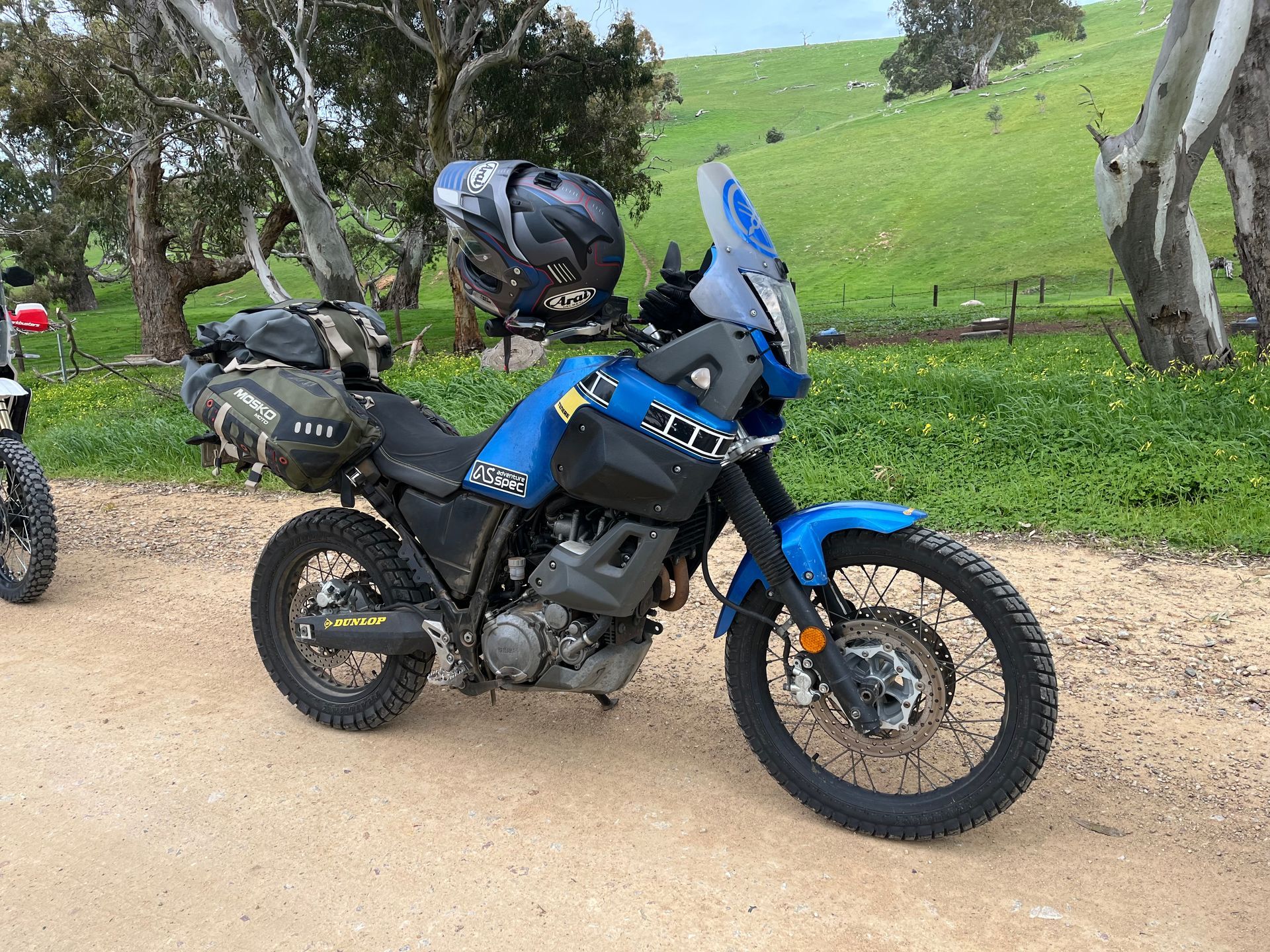 Blue adventure motorcycle parked on a dirt road beside a grassy hillside, with helmet and luggage attached