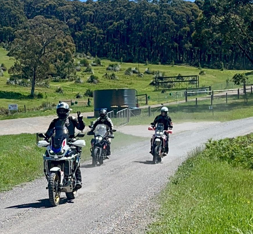 Four motorcyclists ride on a gravel road through a green rural landscape.