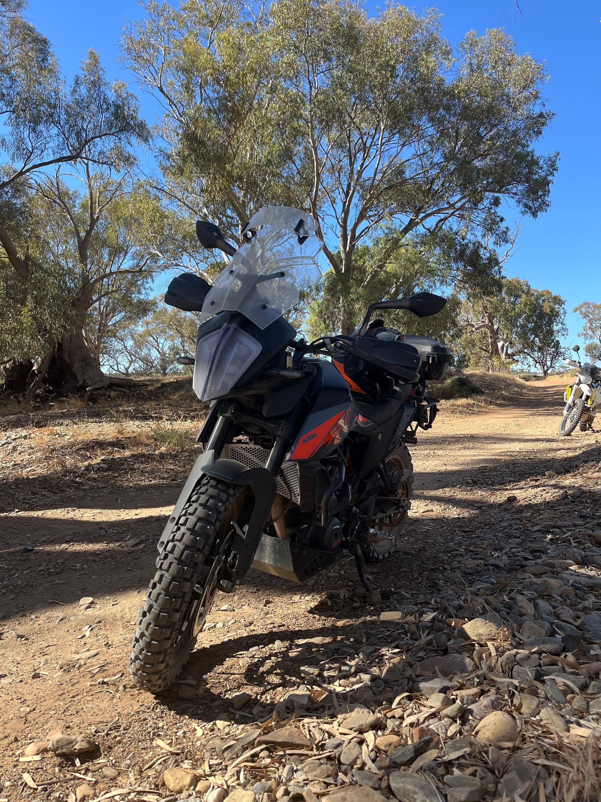 Motorcycle parked on a rocky trail under trees, with a rider standing nearby in the background.
