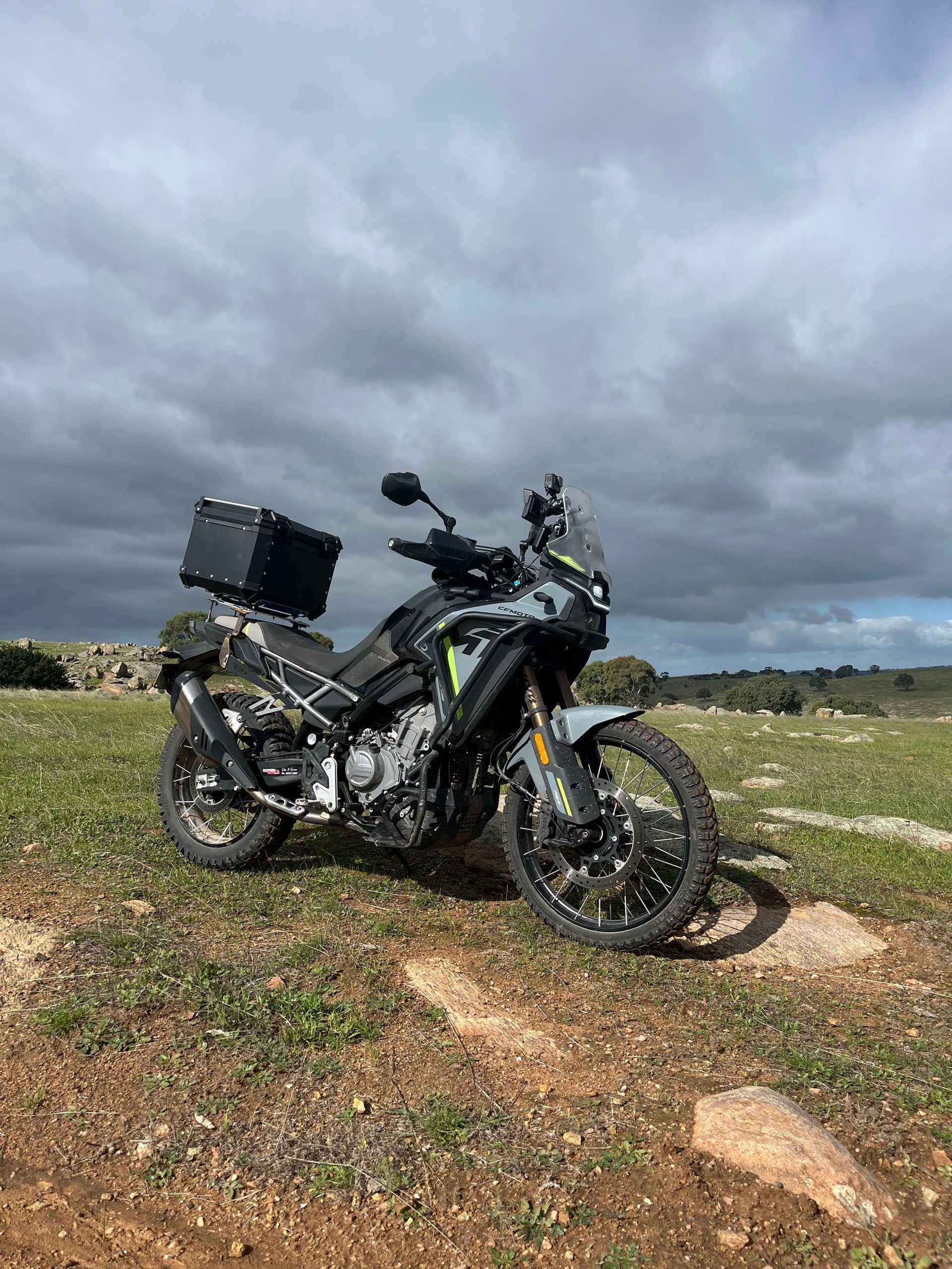 Black adventure motorcycle with top case parked on rocky ground under a cloudy sky.