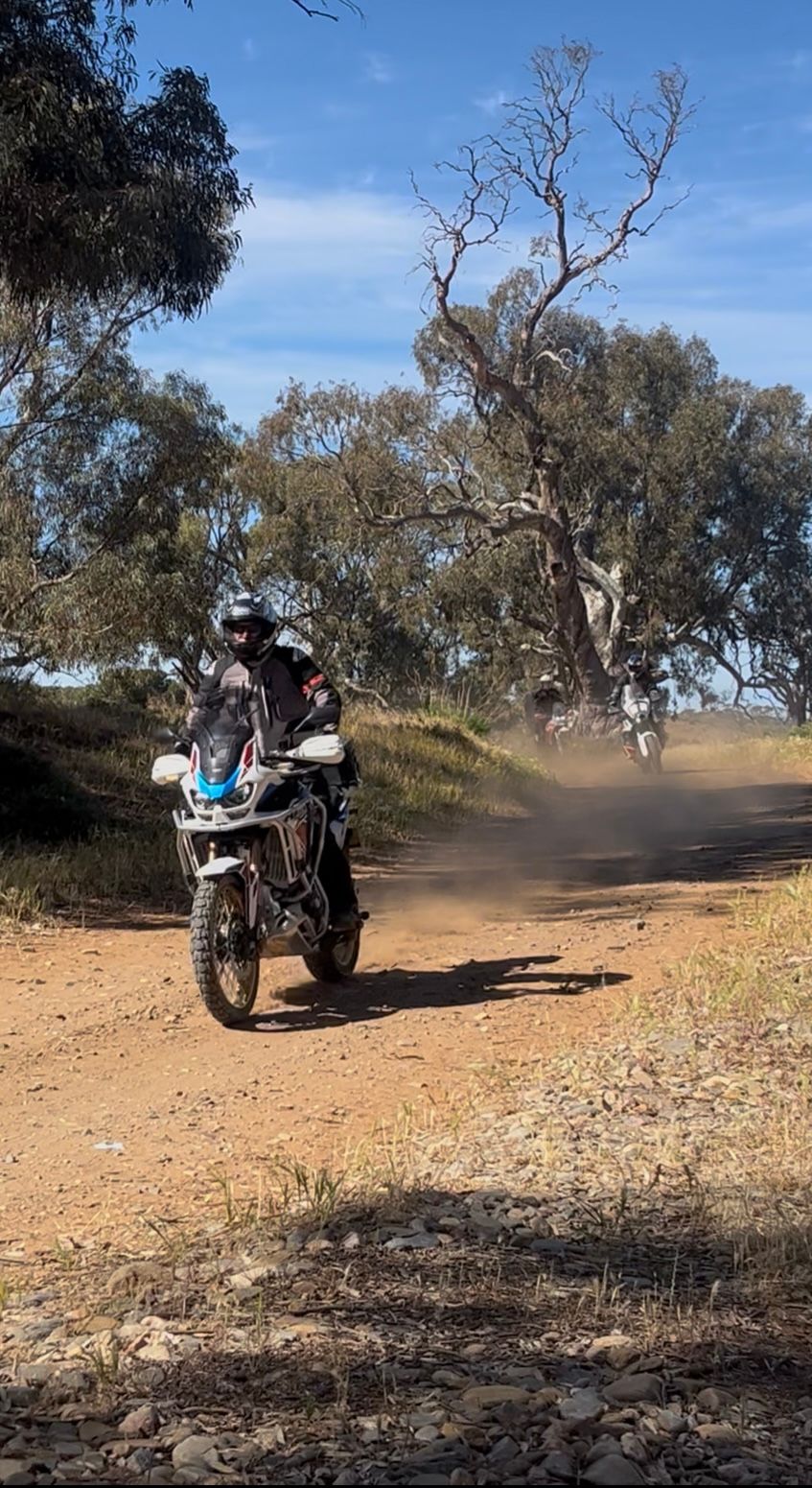 Motorcyclist riding on a dusty dirt trail through a wooded area, with another rider in the distance.