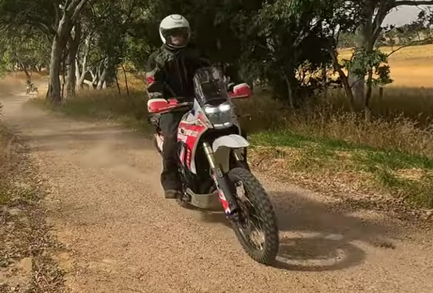 Motorcyclist riding a red-and-white bike on a dusty dirt road lined with trees