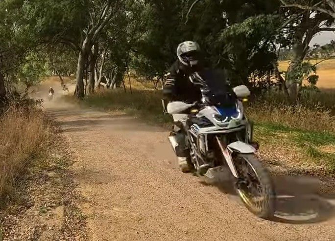 Motorcyclist riding on a dirt path through trees, with dust trailing behind.