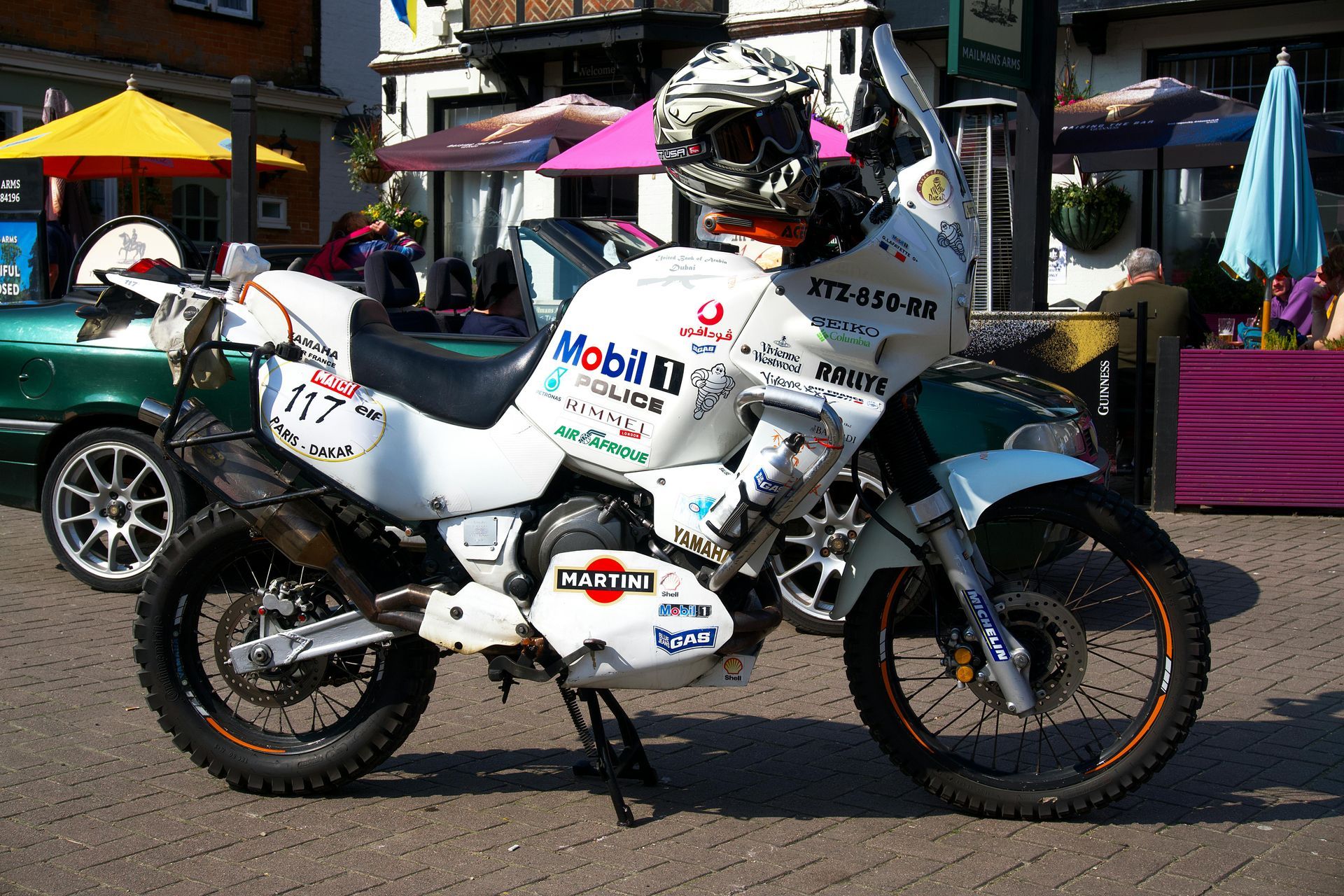 White and blue adventure motorcycle with sponsor logos parked on a sunny street market
