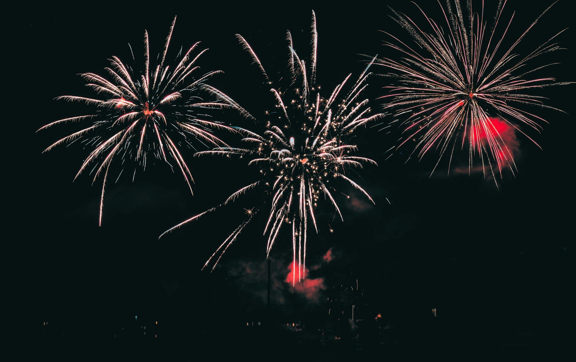 Fireworks bursting in a dark night sky, with red and white sparks over a silhouetted skyline.