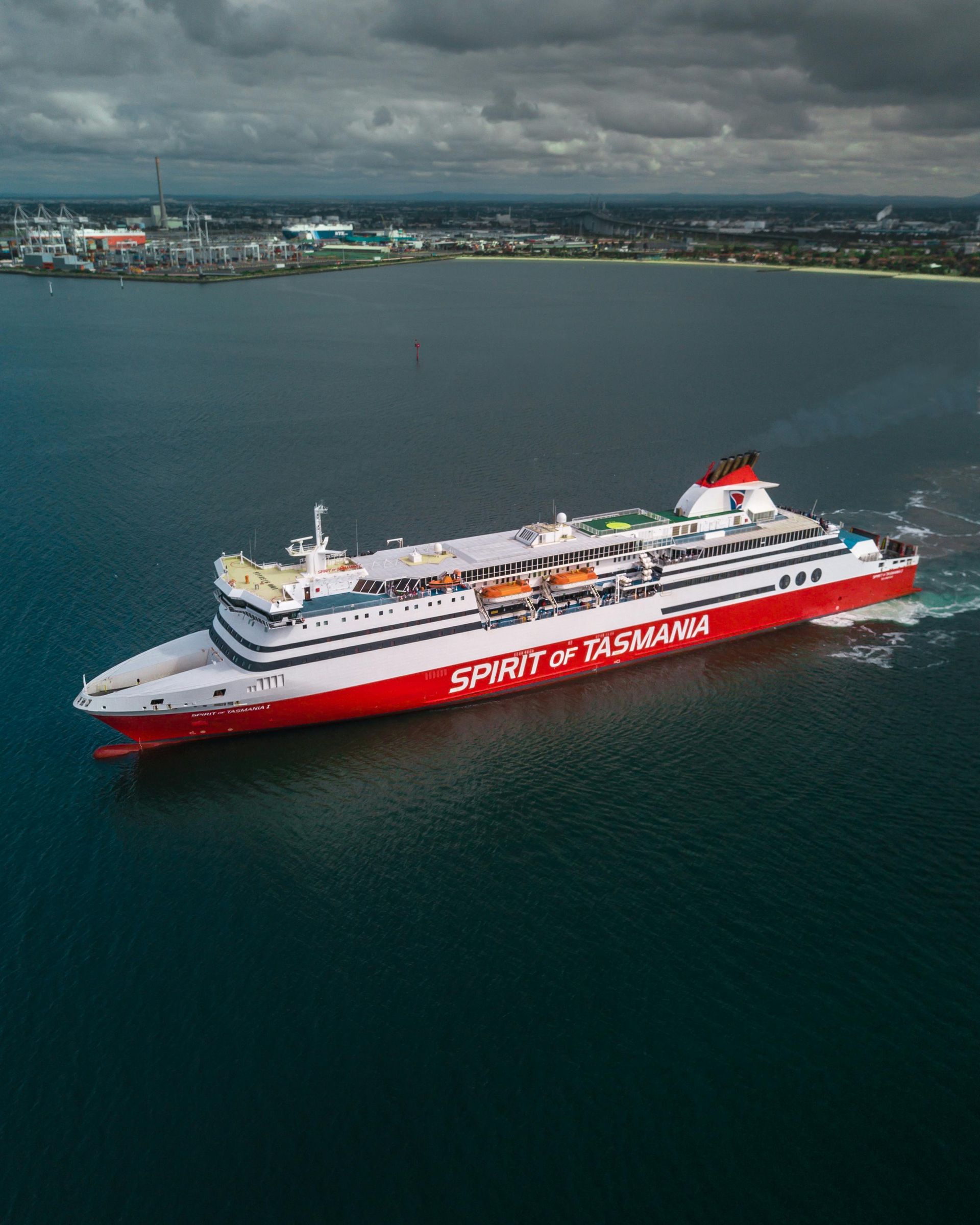 Red and white Spirit of Tasmania ferry sailing on calm water near a coastline under cloudy skies
