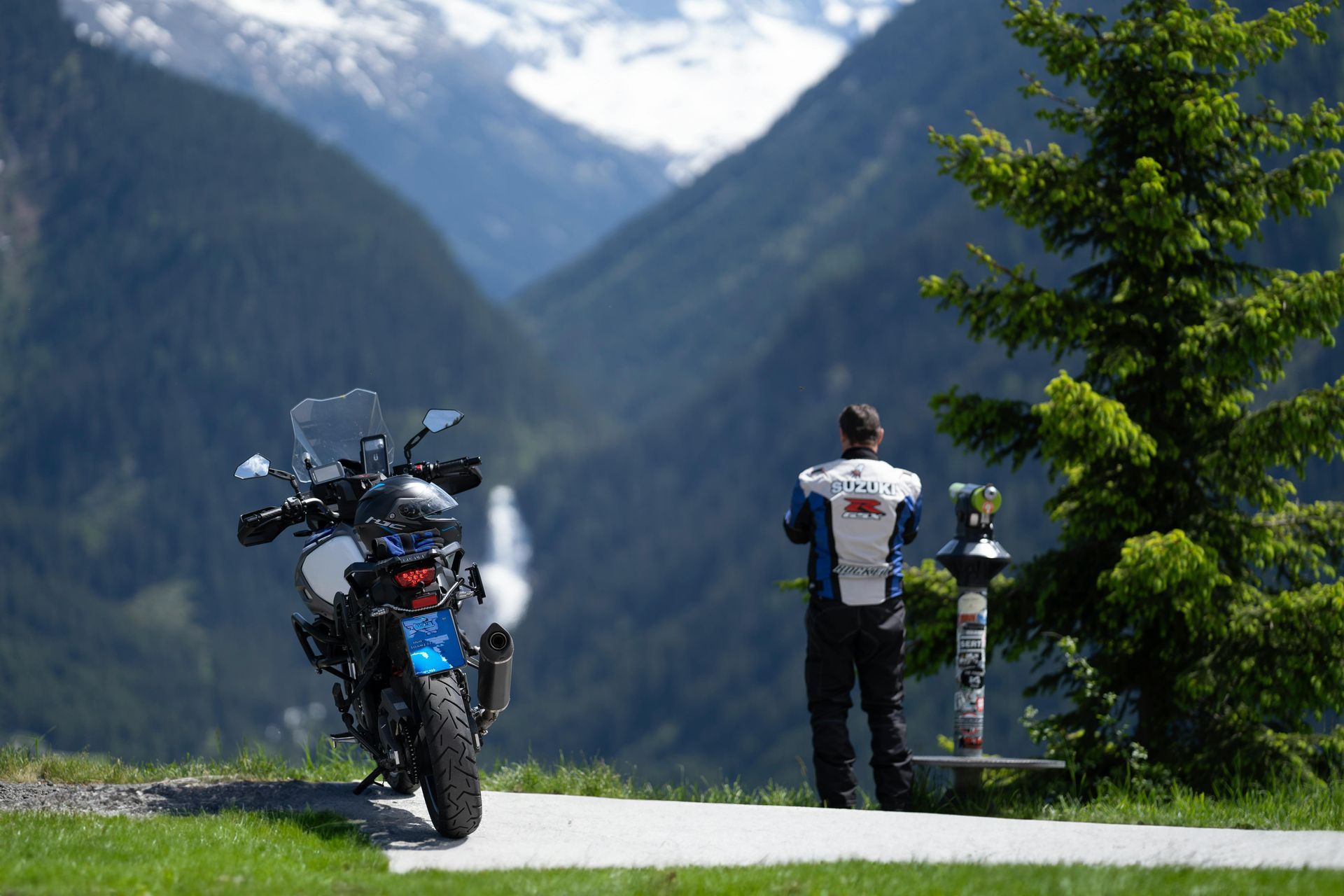 Motorcycle parked on a mountain road beside a person looking over a scenic valley with tall evergreens.
