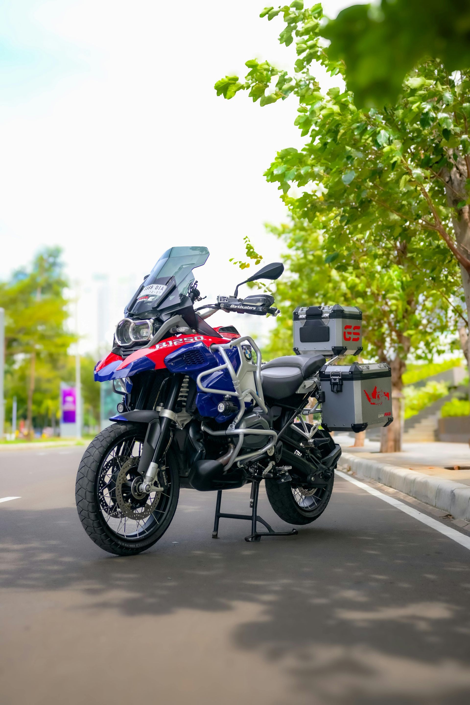 Blue adventure motorcycle parked on a road beside a tree-lined street, with panniers and windshield