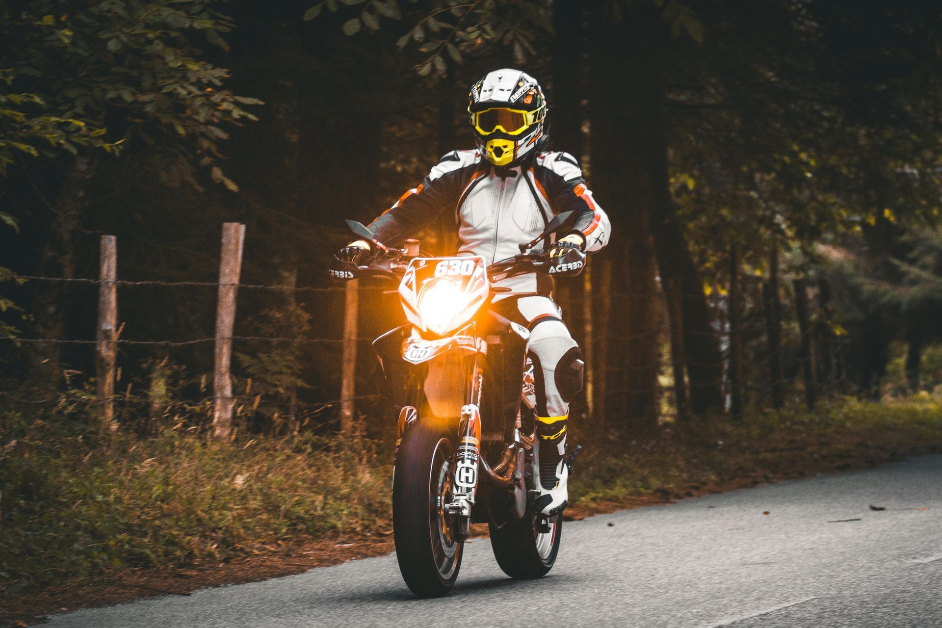 Motorcyclist in white gear riding on a forest road at dusk, headlight shining brightly