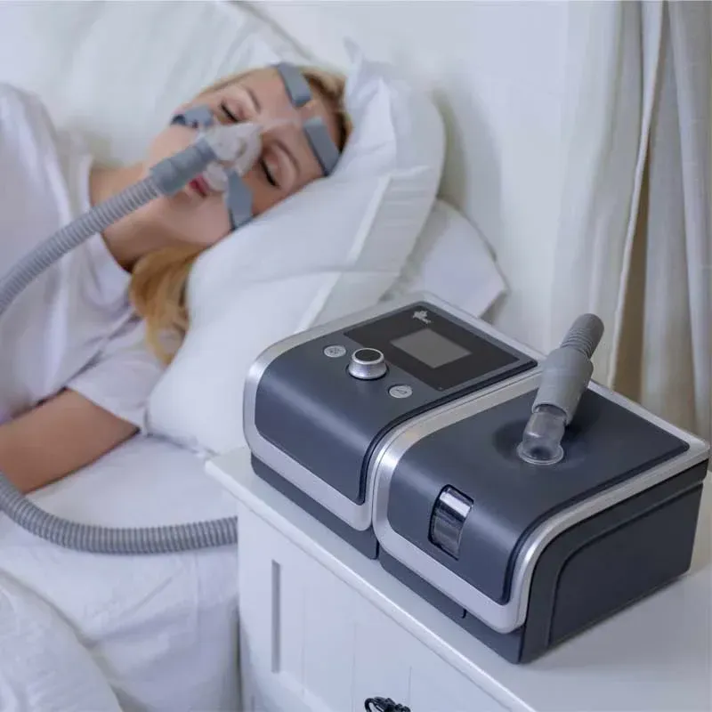 A woman is laying in bed with an oxygen mask on her face and a CPAP machine on her nightstand