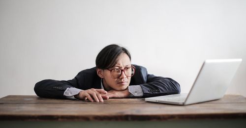 A tired man in a suit and glasses is leaning on a wooden desk in front of a laptop computer.