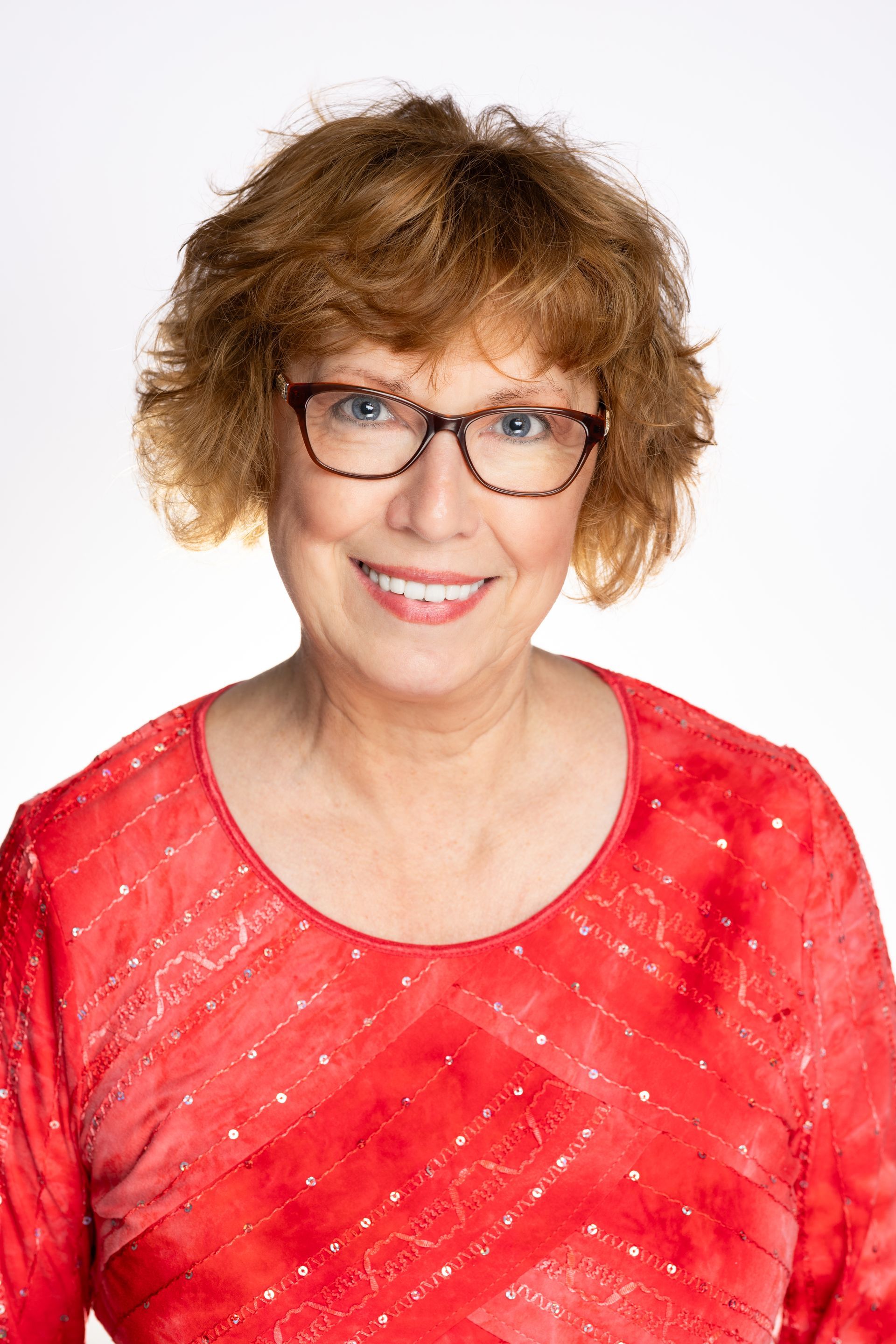 Woman with short, curly auburn hair, wearing glasses and a red patterned top, smiles at the camera.