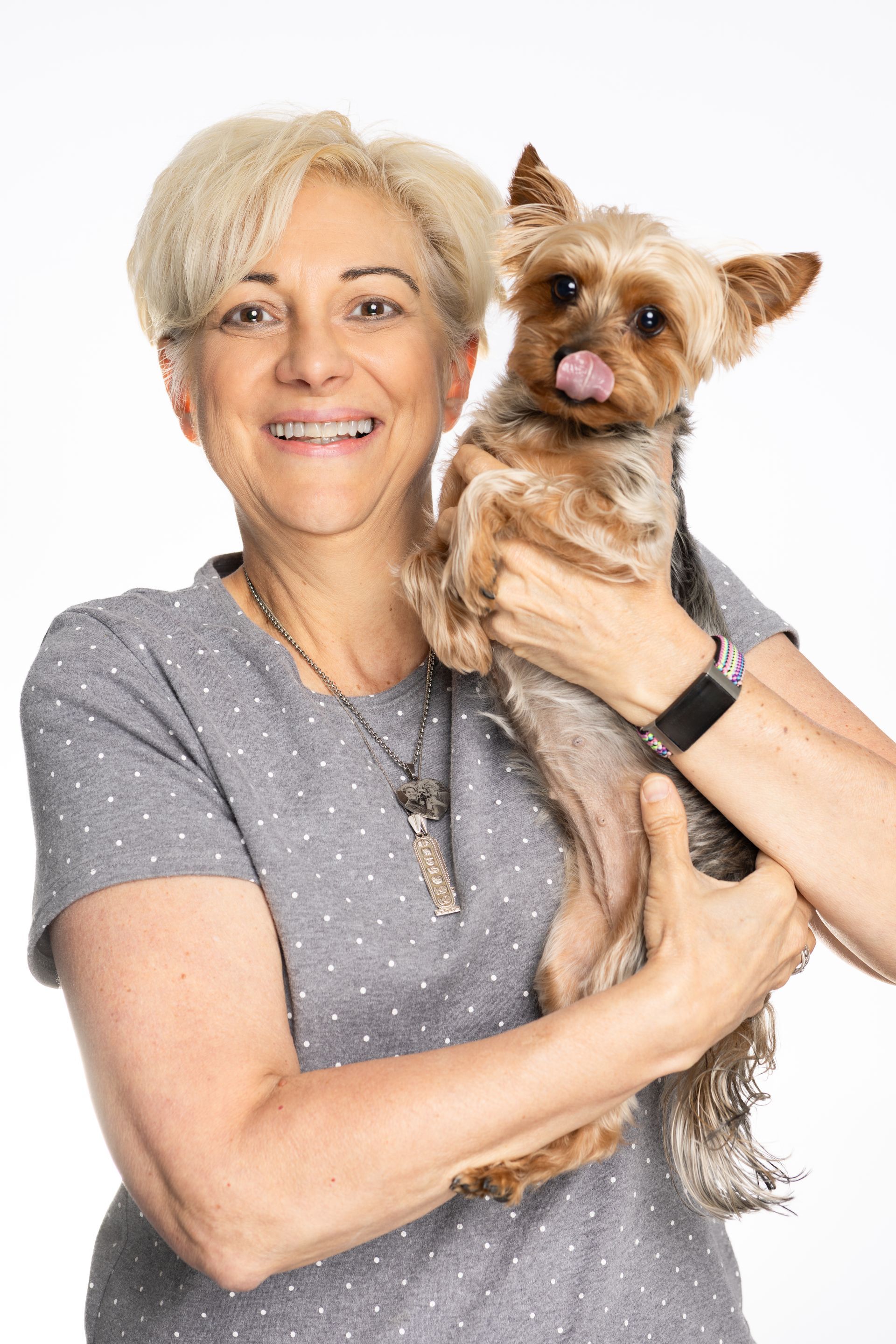 Woman smiling, holding a small dog with tongue out, against a white background.