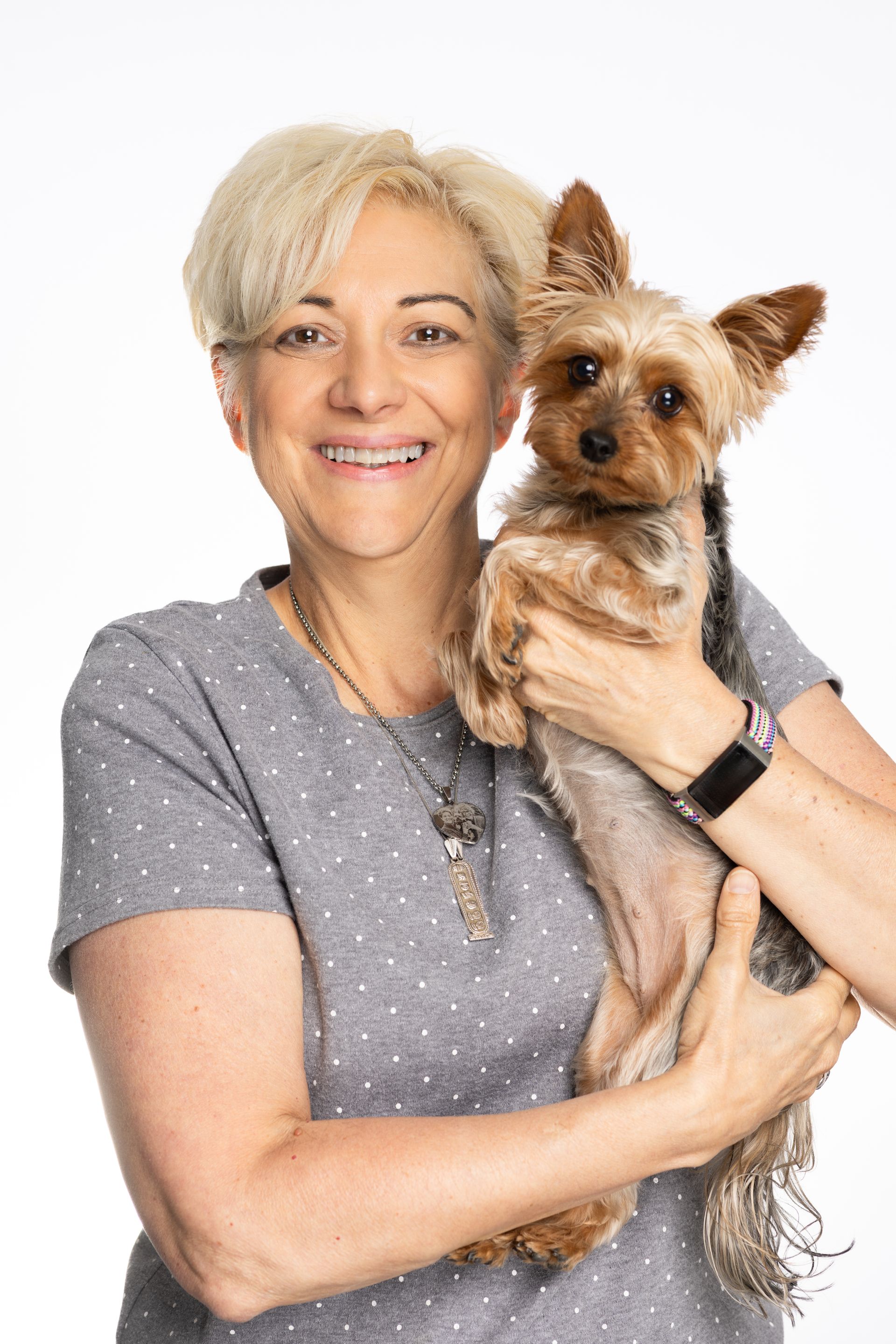 Woman with short blonde hair holds a small Yorkie dog, both smiling, against a white background.