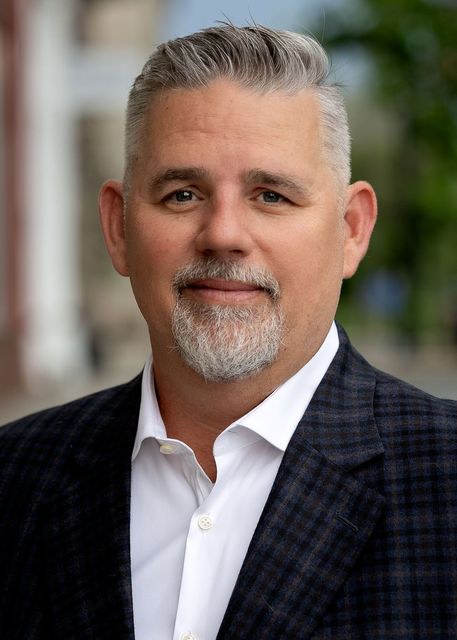 Man in blazer and open-collar shirt smiles, with salt-and-pepper hair and beard, outdoors in front of a building.