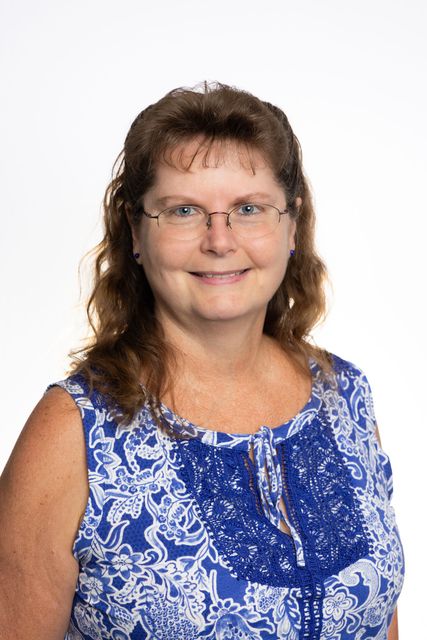 Woman wearing glasses, a blue patterned top, smiling at the camera, against a white backdrop.