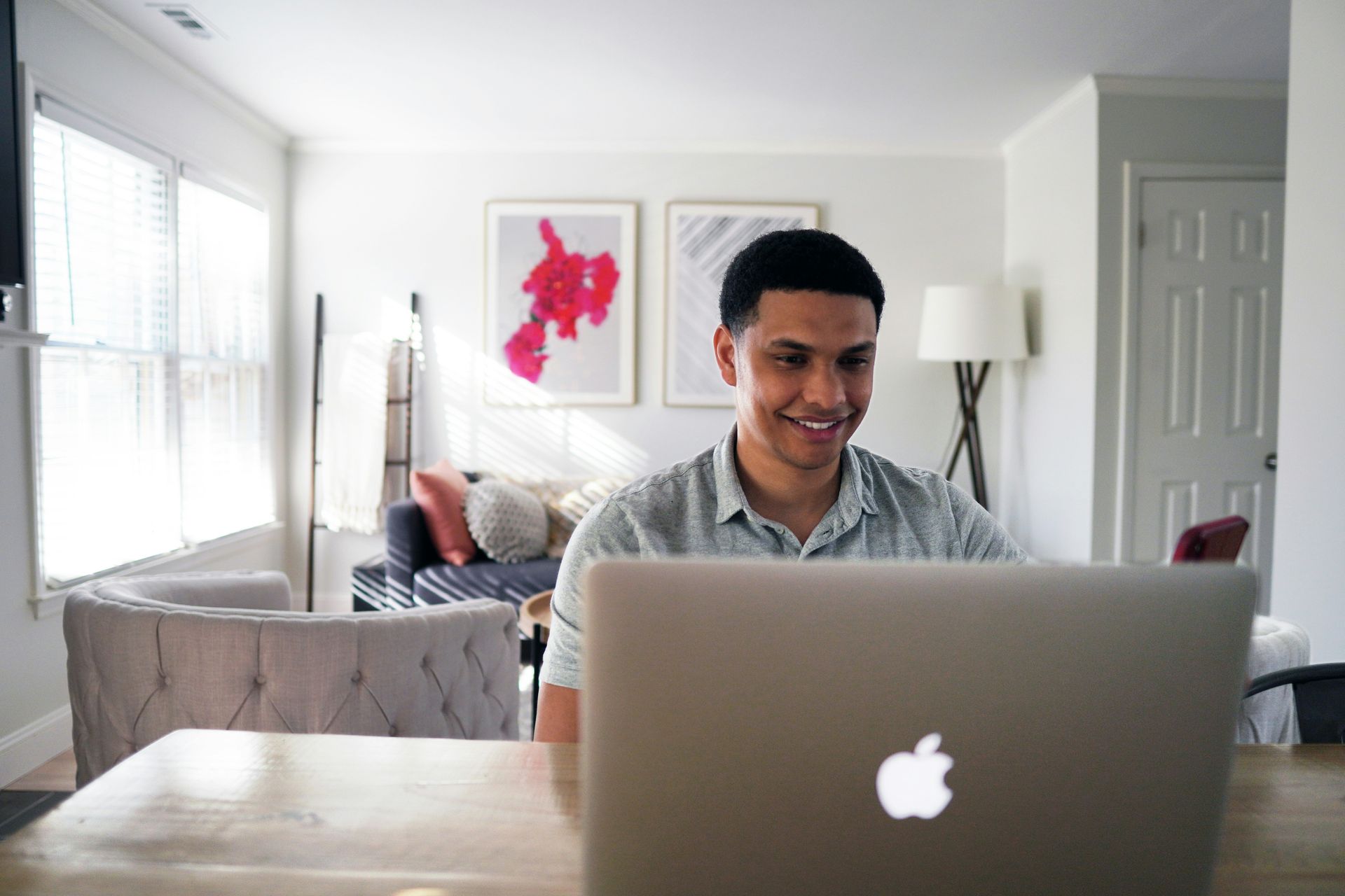 Man smiles while using a laptop at a table in a bright, modern living room.