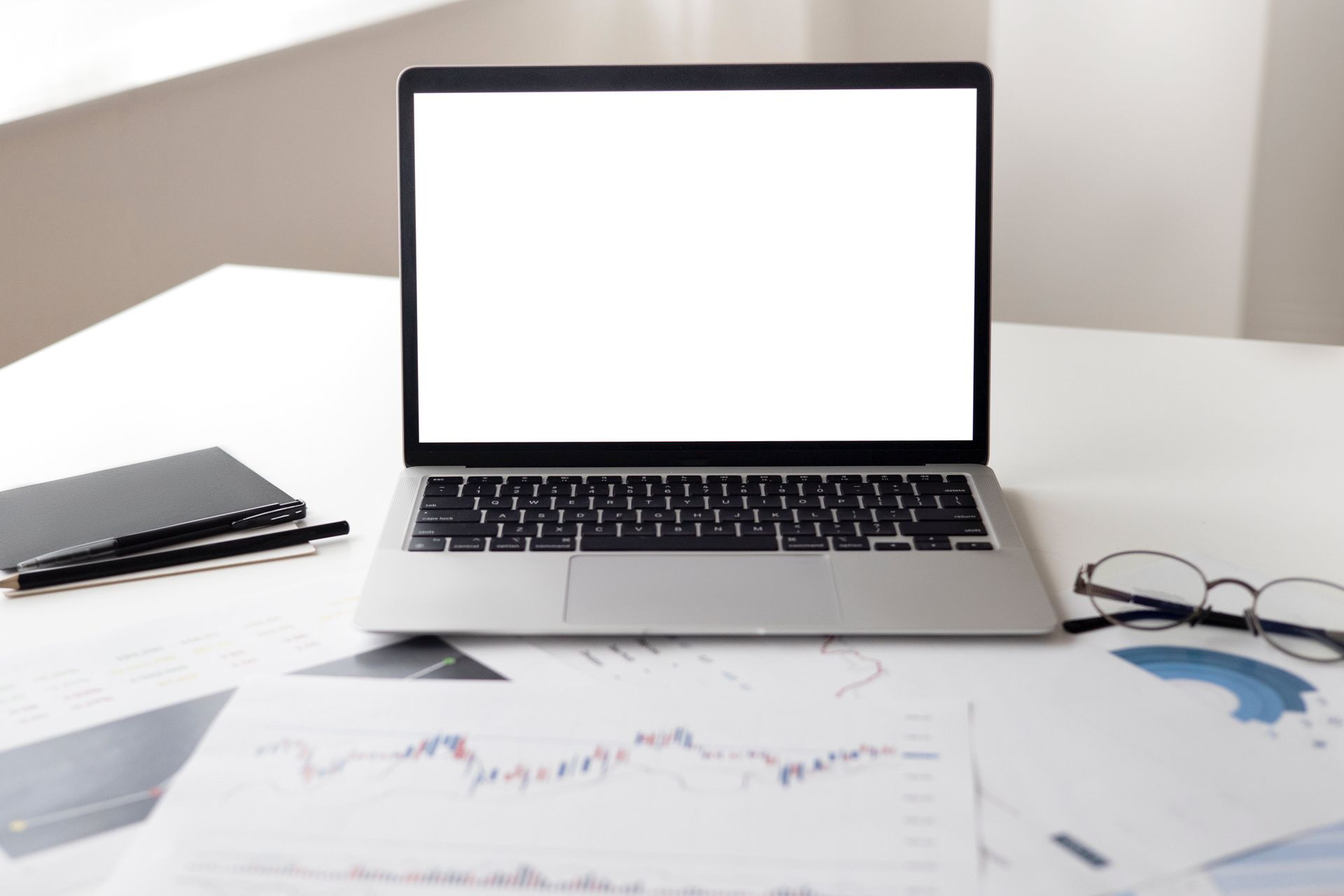Laptop with blank screen on desk with papers, notebook, and glasses.