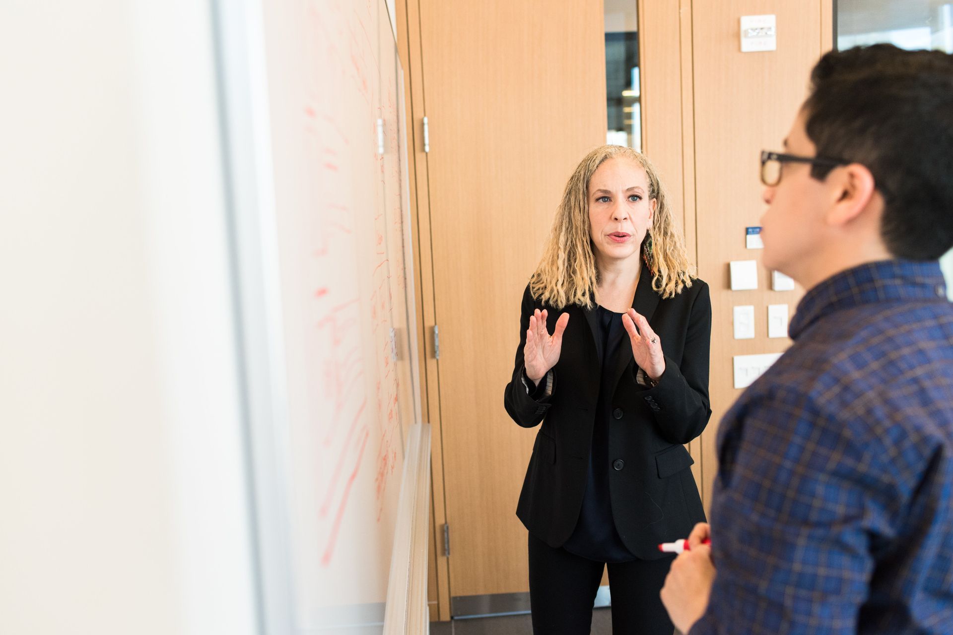 Woman gestures near whiteboard, speaking to person in blue plaid shirt. Office setting.