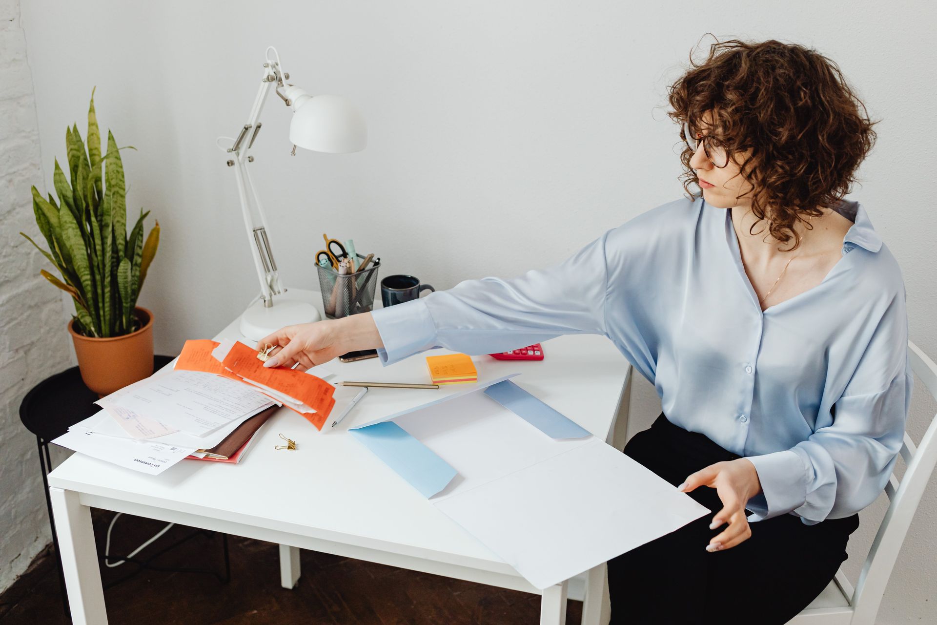 Woman in blue shirt working at white desk with papers, reaching.