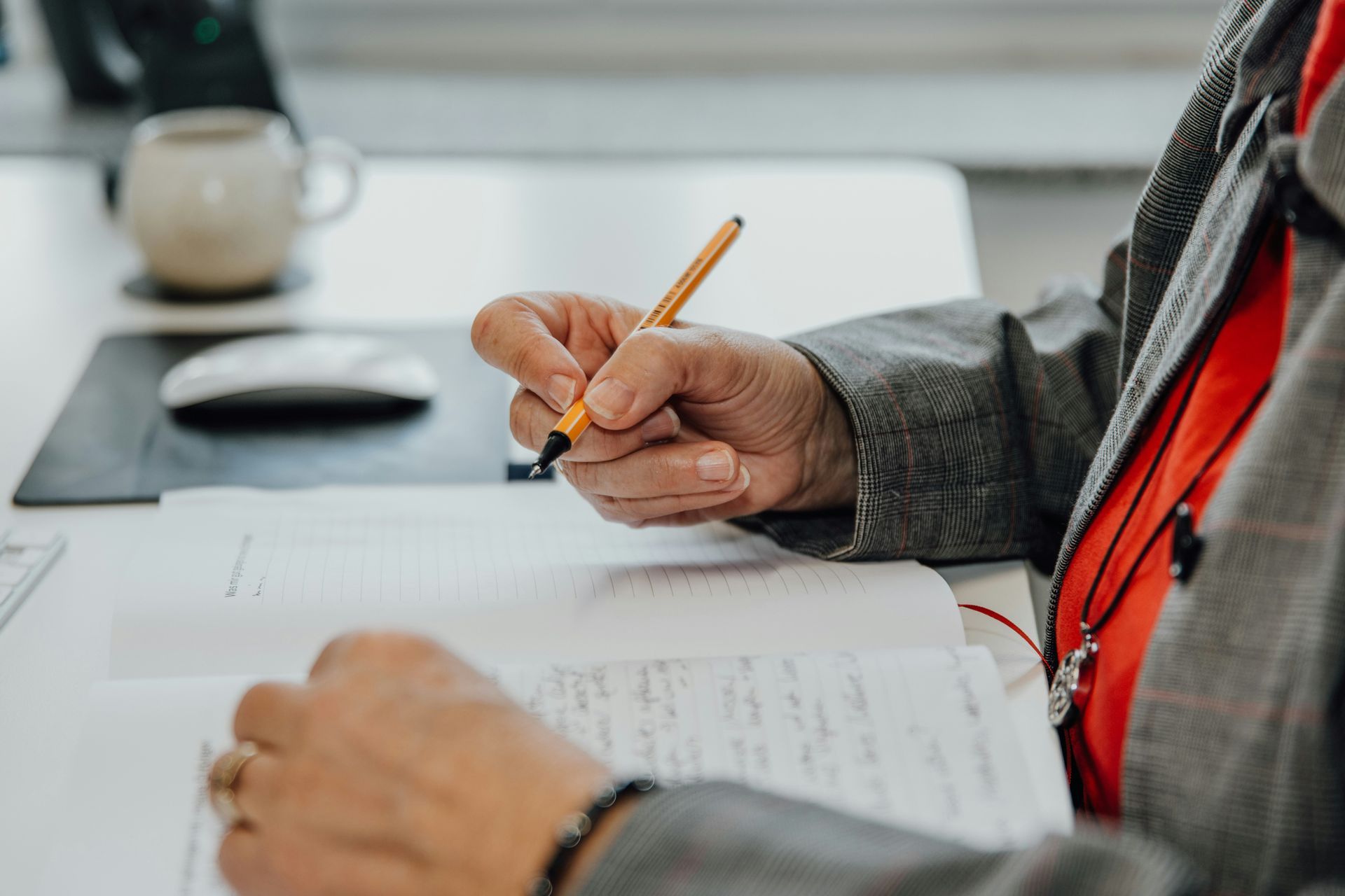Person writing with a pen in a notebook at a desk. Coffee mug and mouse are nearby.