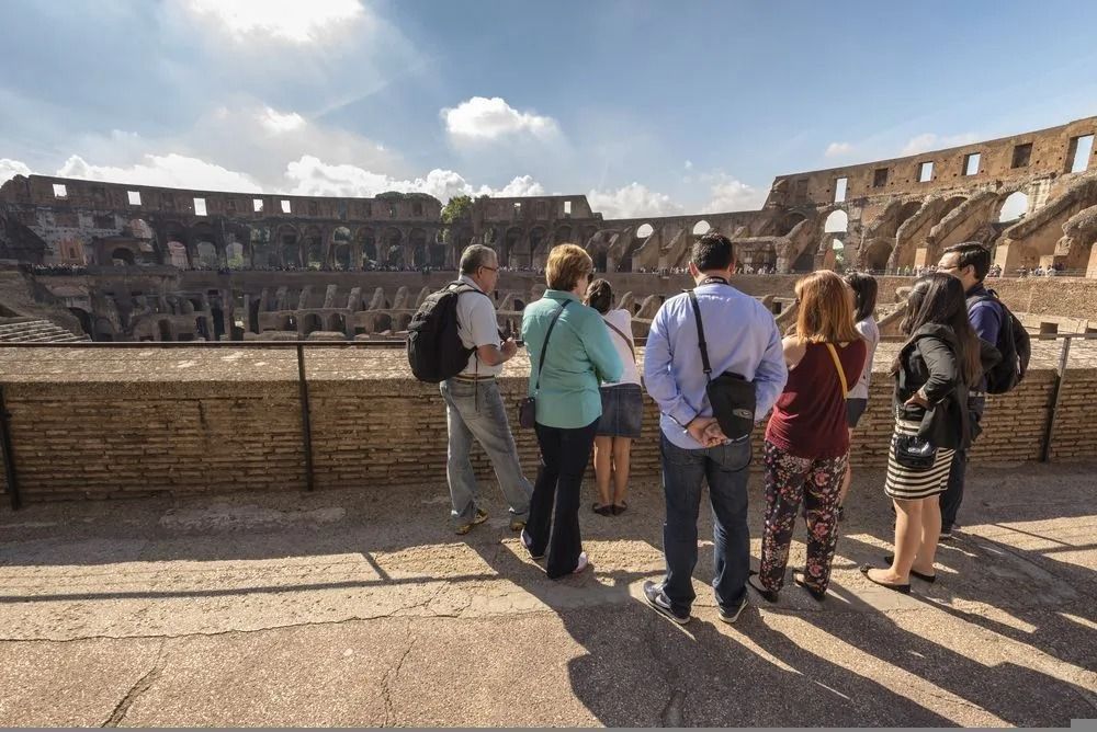 Tourists standing at the Colosseum overlook, with ancient stone arches in the background and sunny skies above