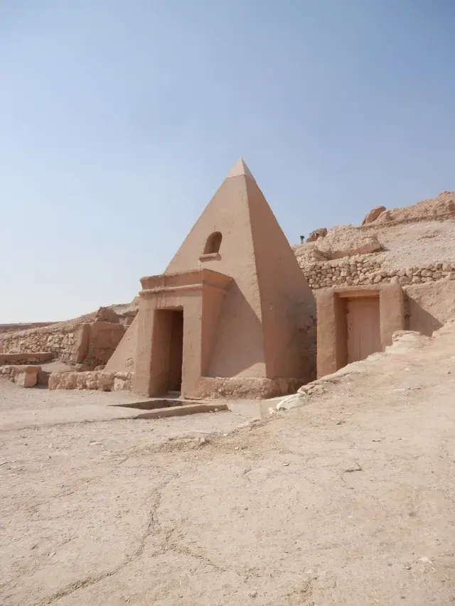 Small desert pyramid-shaped tomb built into rocky cliffs under a clear blue sky.