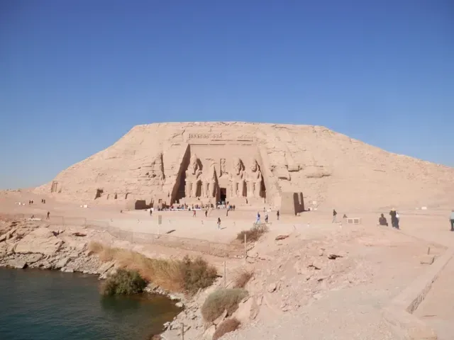 Ancient cliffside temple carved into a sandstone hill beside blue water under a clear sky