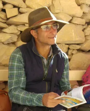 Person in a hat and glasses reading a book outdoors against a stone wall.