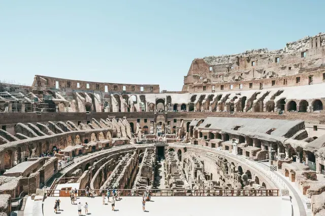 Ancient Roman Colosseum interior with arched seating and visitors under a blue sky