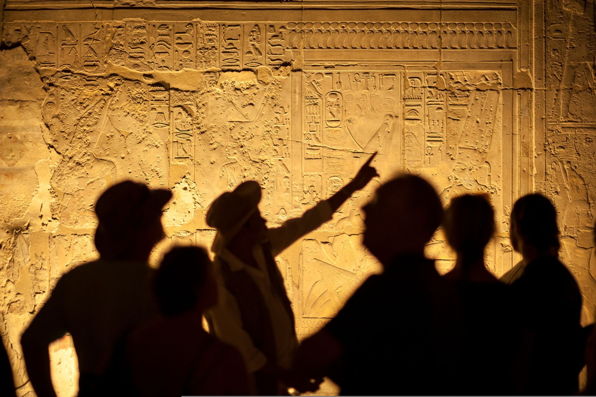 Silhouetted people in front of an illuminated carved wall, one pointing upward.