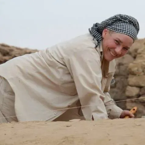 Smiling person in beige clothing crawling on desert sand beside rocks, wearing a checkered headscarf