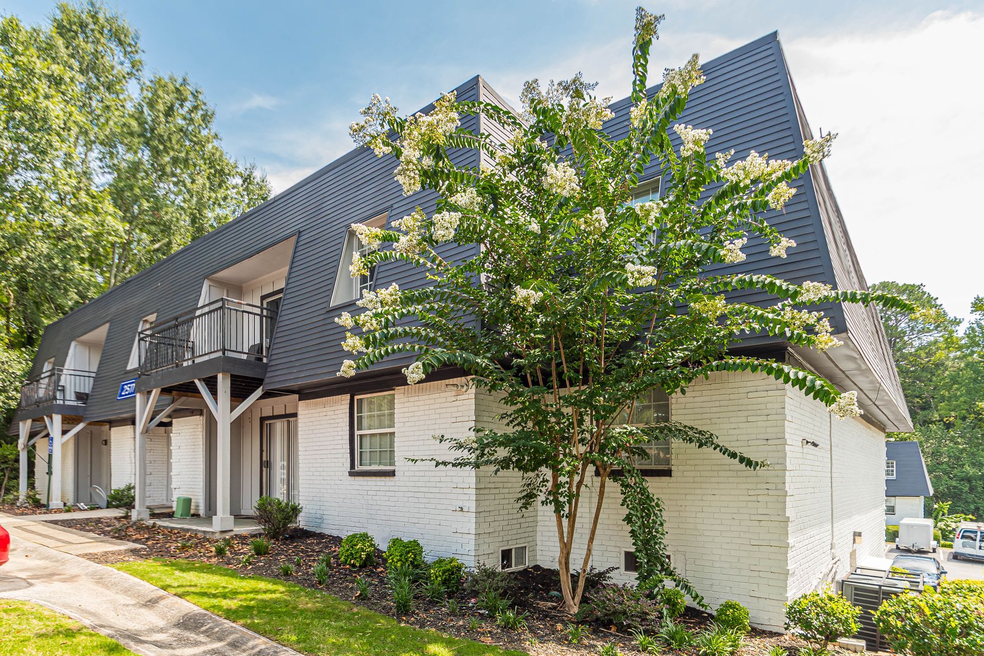 White brick house with dark shingled roof and front porch, surrounded by trees and landscaping