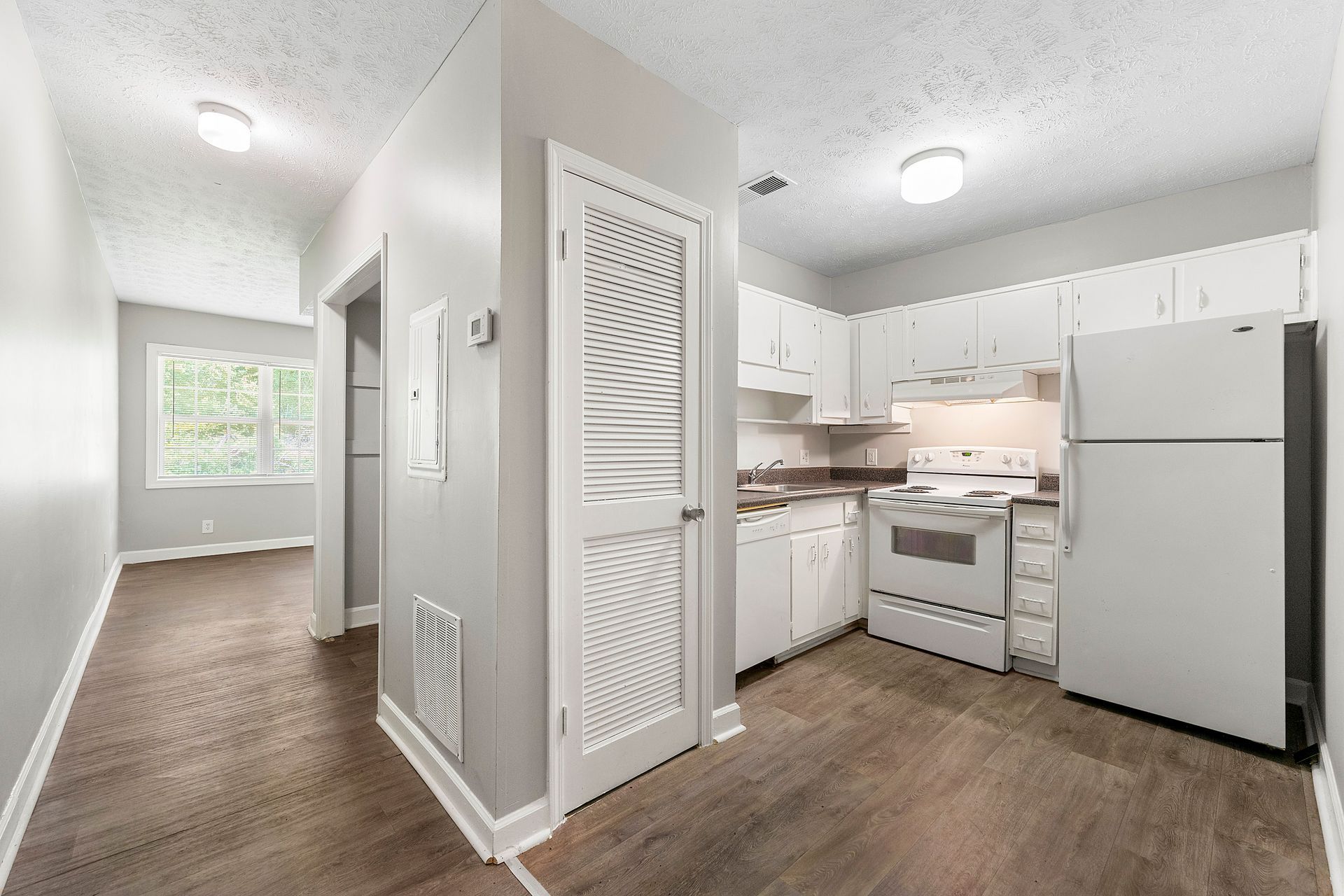 Bright white kitchen with stove, fridge, and cabinets, opening to a hallway with a window and wood flooring.