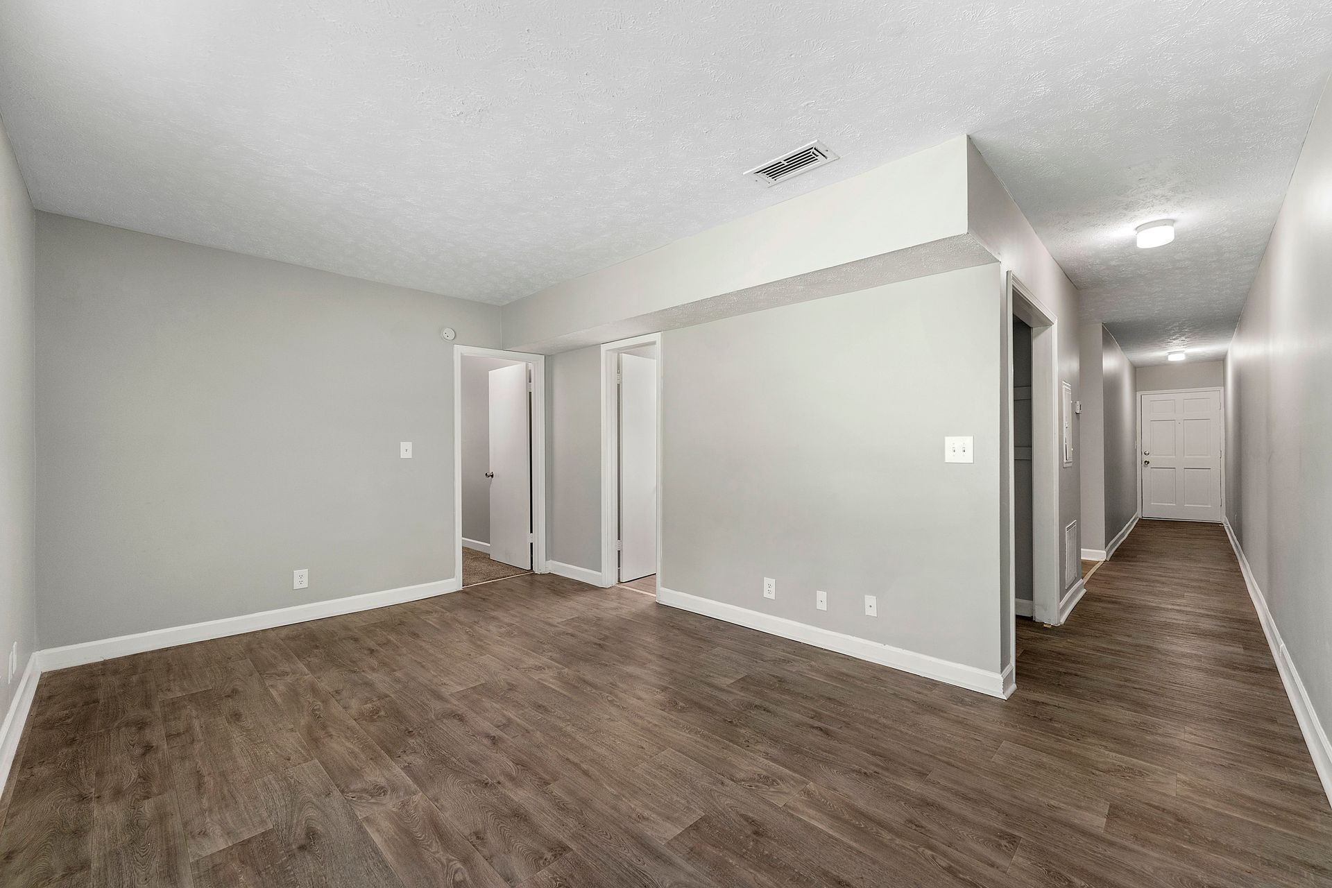 Empty beige room with dark wood flooring, white trim, and open doorways to a hallway.