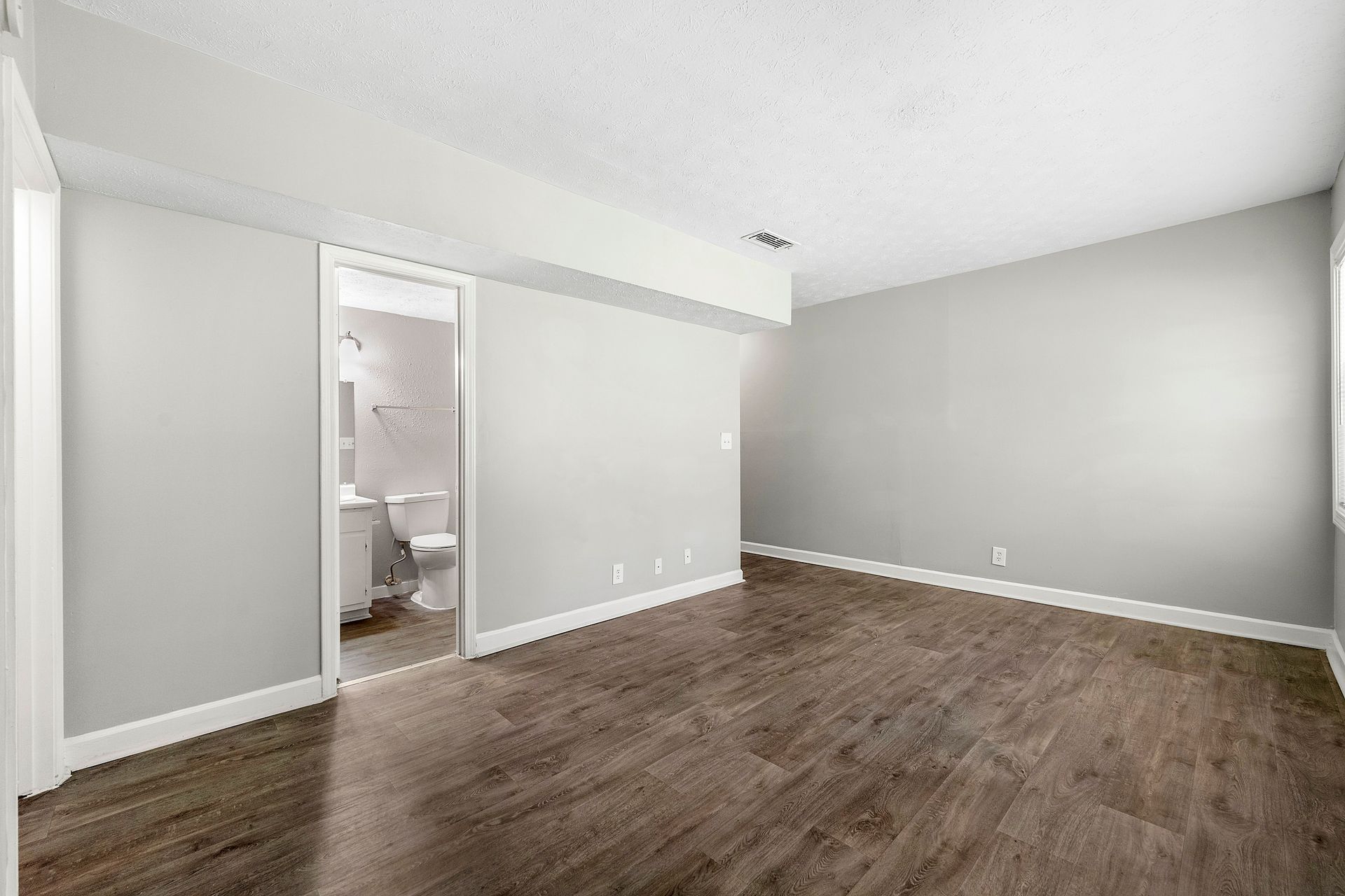 Empty bedroom with gray walls, white trim, and wood flooring, viewed toward an open bathroom doorway