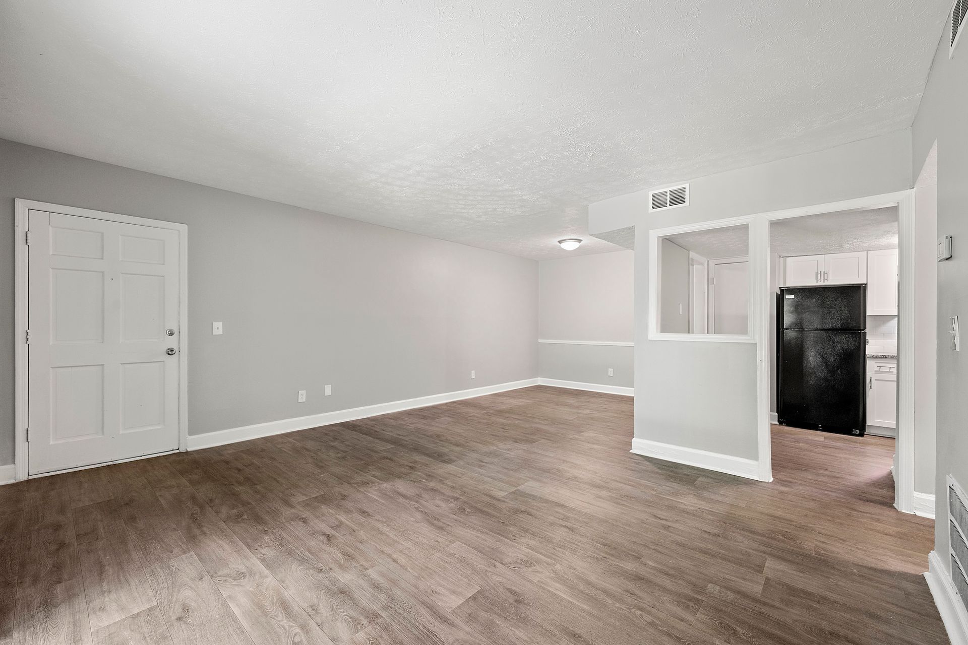 Empty gray living room with wood floors, white trim, and a small pass-through opening to a kitchen area