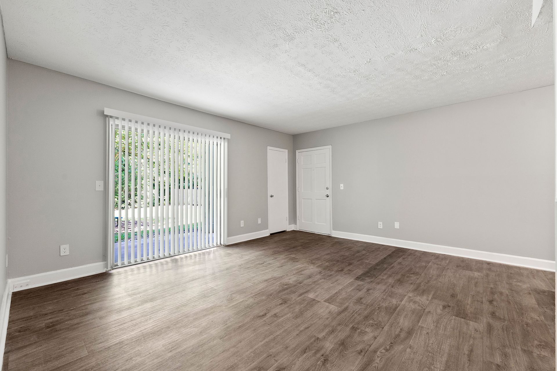 Empty beige living room with brown carpet, white walls, and sliding glass door with vertical blinds