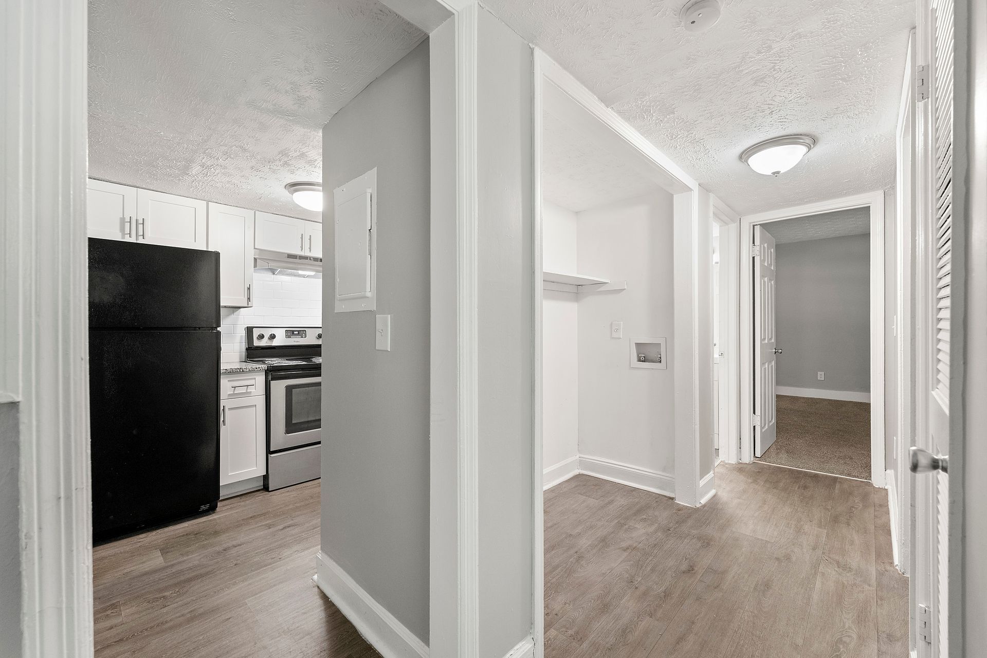 Bright apartment hallway with wood floors, white walls, and a view into the kitchen and entryway