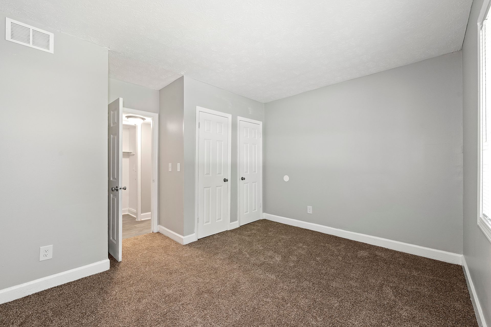 Empty carpeted bedroom with light gray walls, white closet doors, and a window on the right.