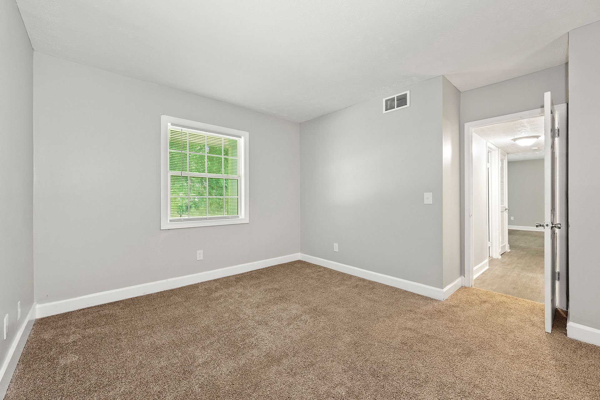 Empty beige-carpeted bedroom with gray walls, a window, and an open doorway to a hall.
