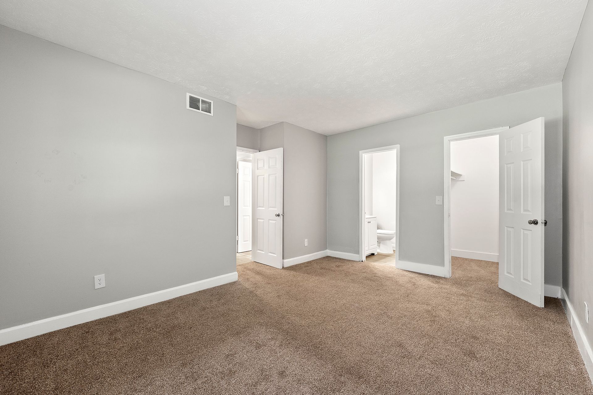 Empty beige-carpeted bedroom with gray walls and two open doorways to a bathroom and hall.