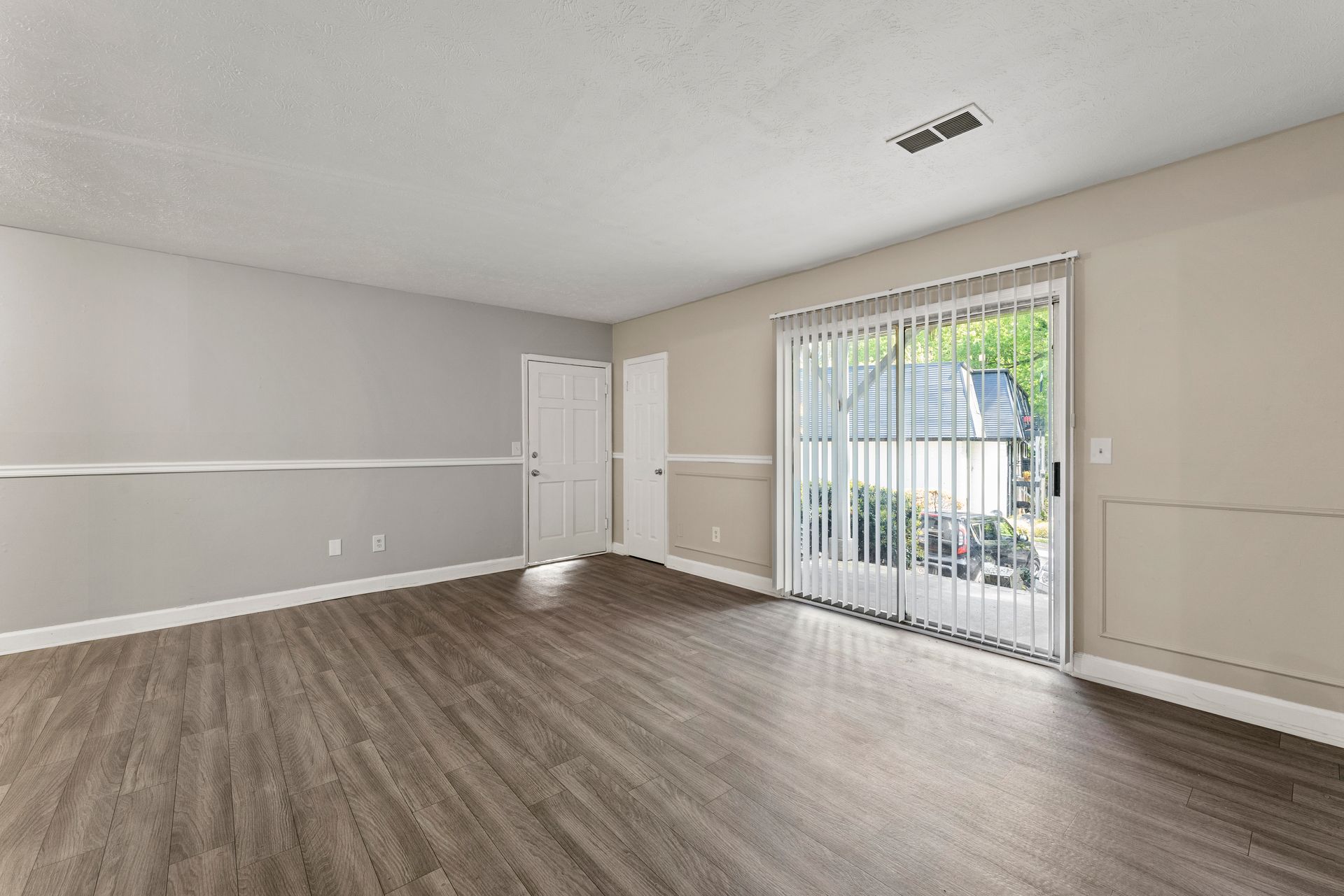 Empty living room with gray walls, wood-look flooring, and a sliding glass door with vertical blinds.