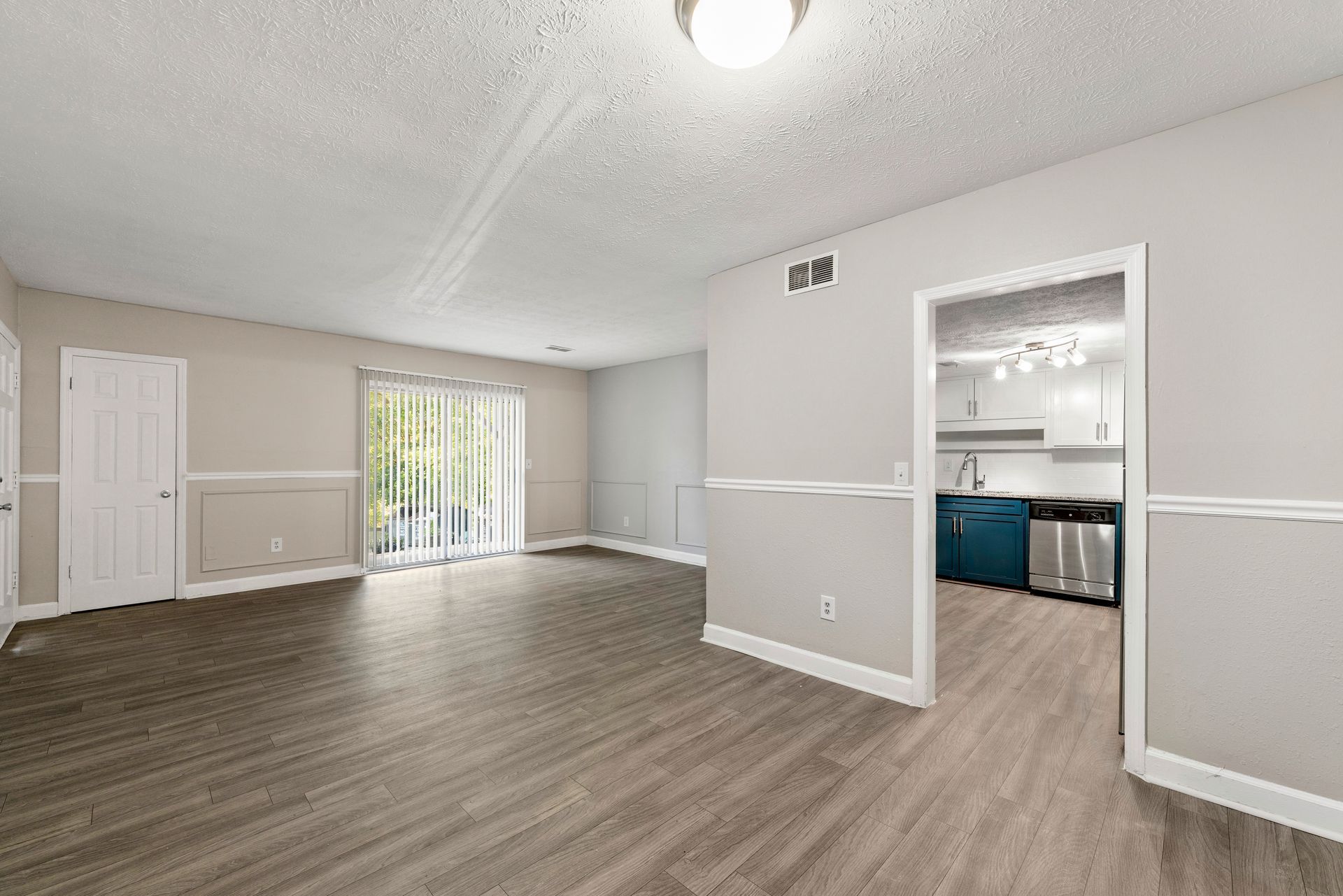 Empty open-plan room with gray flooring and light walls, leading into a kitchen with blue cabinets.