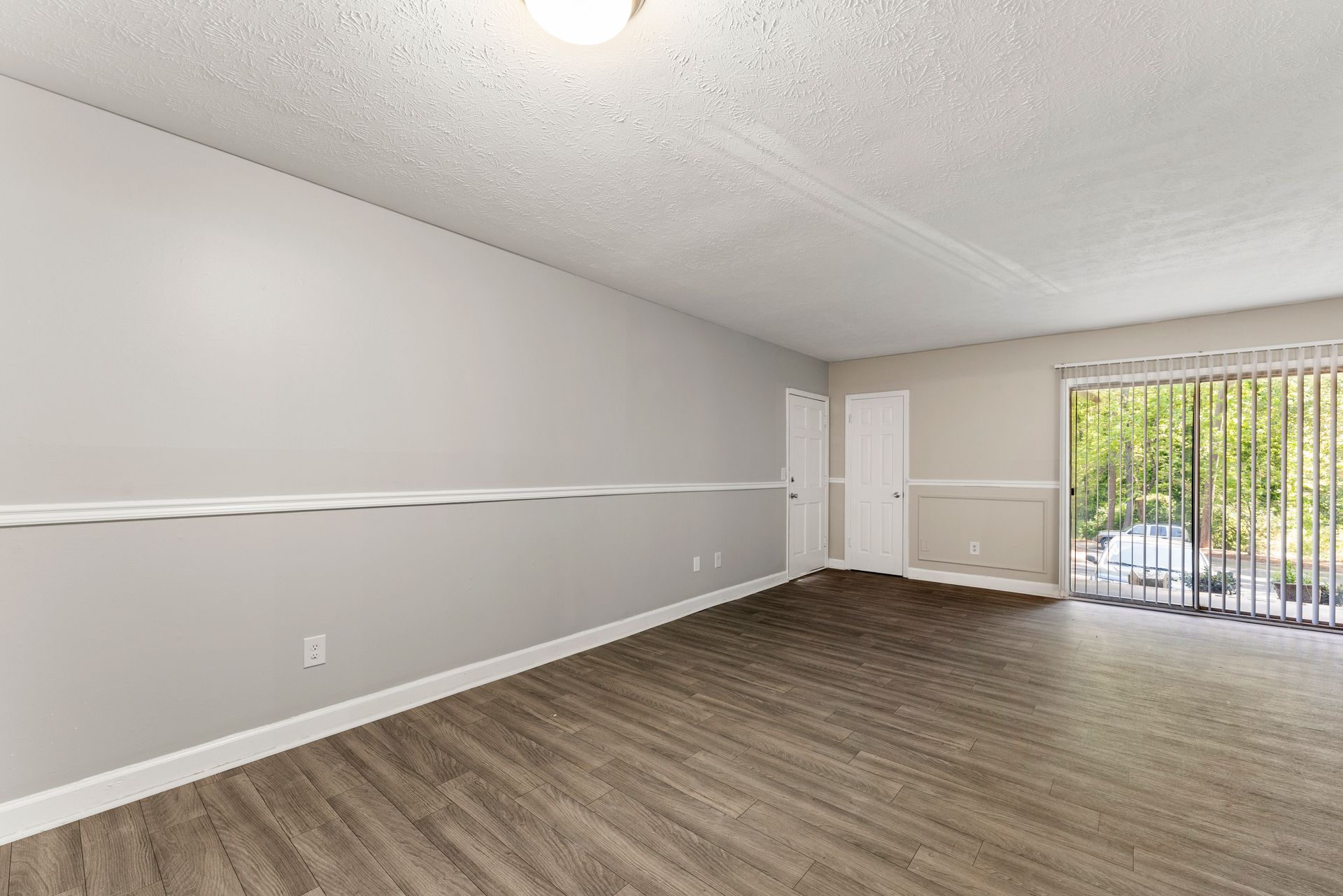 Empty living room with gray walls, wood flooring, and sliding glass door with vertical blinds