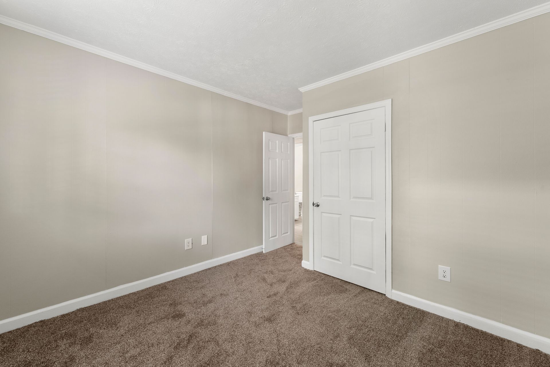 Empty beige bedroom with carpet, white trim, open door, and closet doors.
