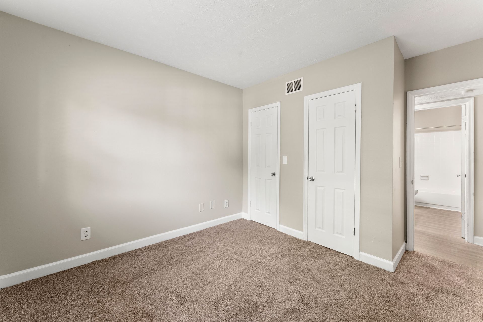 Empty beige bedroom with carpet, white doors, and an open doorway to a bright hall