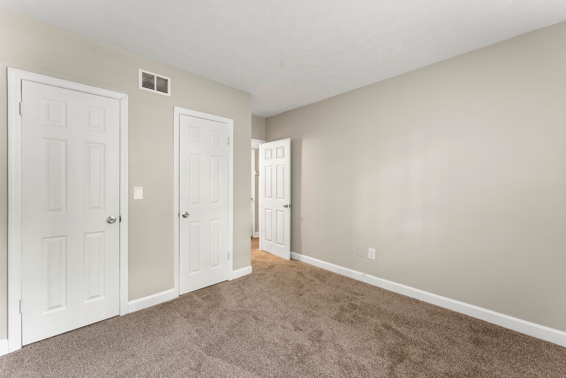Empty beige bedroom with carpet, two white closets, and an open doorway.